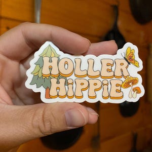 May include: A white sticker with the text "Holler Hippie" in orange and yellow letters. The sticker features a green pine tree, a butterfly, and mushrooms.