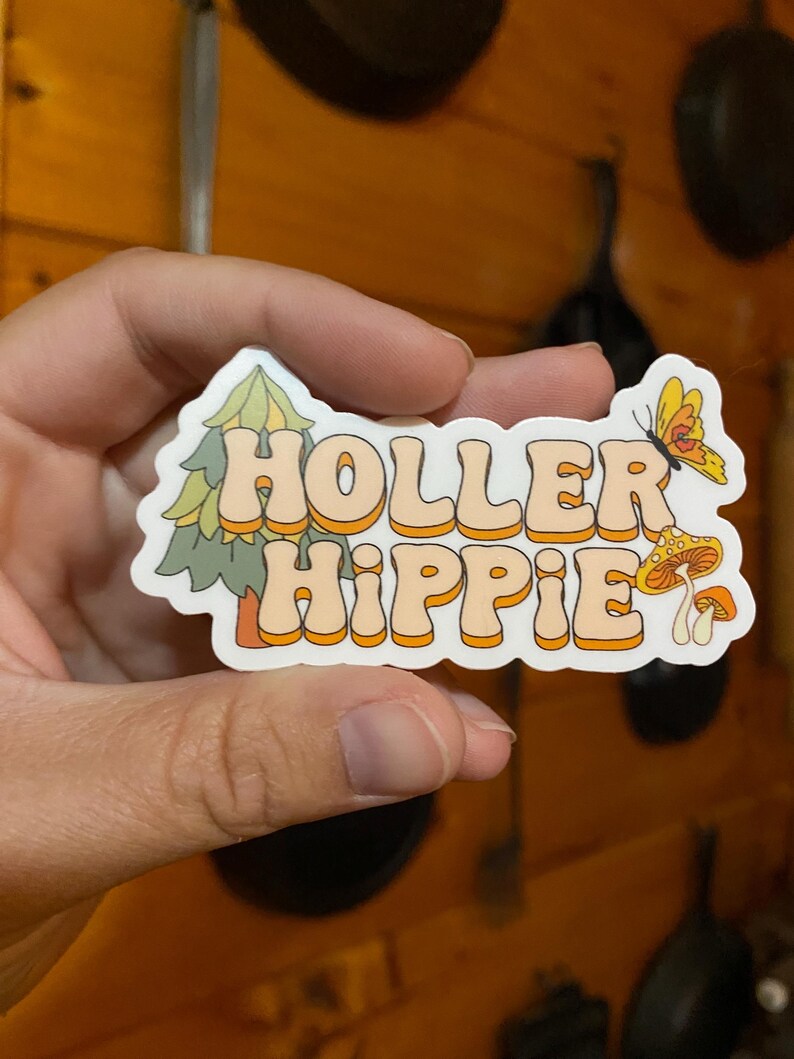 May include: A white sticker with the text "Holler Hippie" in orange and yellow letters. The sticker features a green pine tree, a butterfly, and mushrooms.