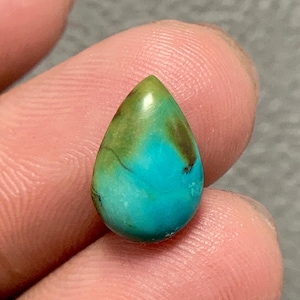 May include: A teardrop-shaped turquoise gemstone with a polished surface. The stone features a vibrant mix of colors, including shades of turquoise, green, and brown. The gemstone is held between fingers, showcasing its size and shape.