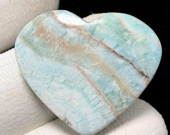 May include: A heart-shaped gemstone with a polished surface. The stone displays a mix of light blue, beige, and brown colours, with a striped pattern. The gemstone is held by a white gloved hand, set against a black background.