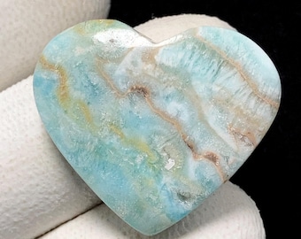 May include: A heart-shaped gemstone with a polished surface. The stone displays a mix of light blue, white, and tan hues, with natural veining. The gemstone is held in white fabric.
