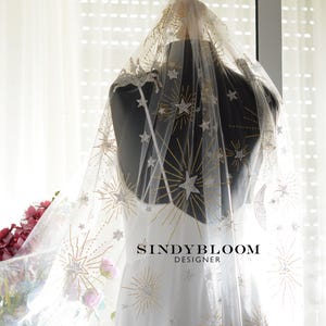 Può includere: Un velo da sposa bianco trasparente con un design celeste. Il velo è adornato con stelle, lune e raggi di sole dorati e argentati. Il velo è drappeggiato su un manichino che indossa un abito bianco. Il nome del designer, SINDYBLOOM DESIGNER, è stampato sull'abito.