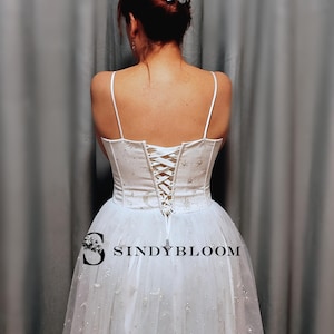 May include: A white corset-style top with thin spaghetti straps and a lace-up back closure, featuring a celestial pattern of silver stars and moons. The garment is paired with a matching white tulle skirt adorned with silver star and moon accents.