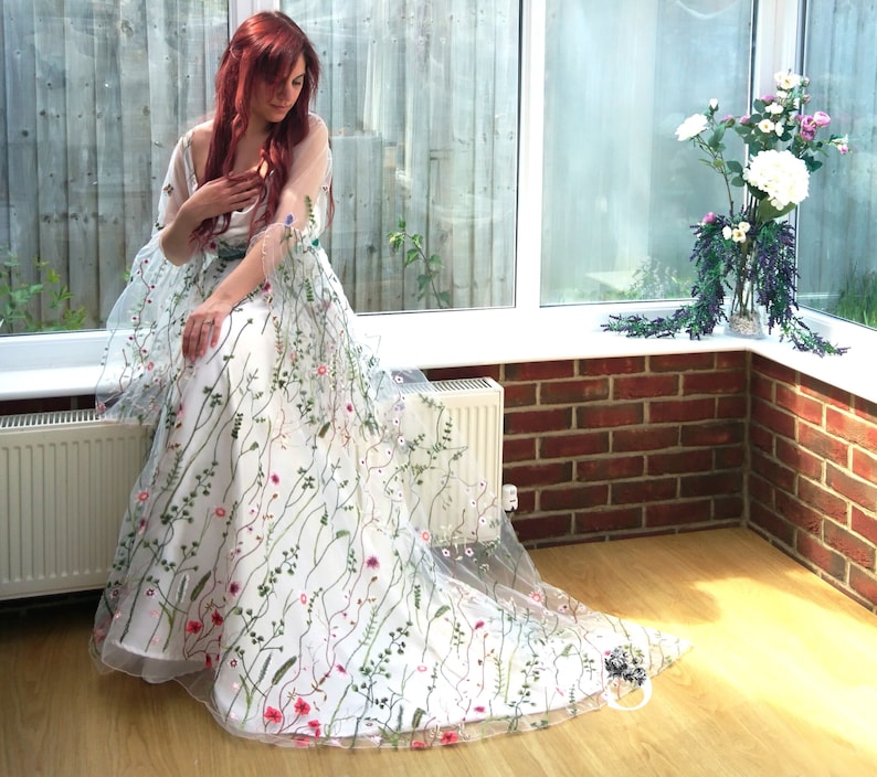 Puede incluir: Un vestido blanco largo hasta el suelo con mangas fluidas y transparentes, cubierto por un delicado bordado floral multicolor. El vestido presenta una variedad de tallos verdes, hojas y peque&ntilde;as flores rosas, rojas y moradas sobre la tela.