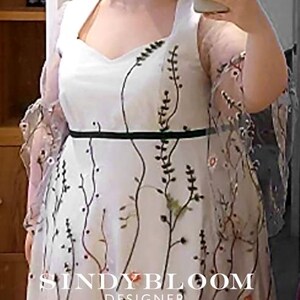 Puede incluir: Un vestido blanco con escote cuadrado, una fina banda negra en la cintura y mangas transparentes. La tela est&aacute; adornada con un patr&oacute;n bordado de enredaderas verticales de color verde oscuro y peque&ntilde;as flores coloridas. El texto SINDY BLOOM DESIGNER es visible.