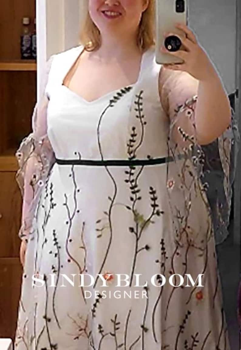 Puede incluir: Un vestido blanco con escote cuadrado, una fina banda negra en la cintura y mangas transparentes. La tela est&aacute; adornada con un patr&oacute;n bordado de enredaderas verticales de color verde oscuro y peque&ntilde;as flores coloridas. El texto SINDY BLOOM DESIGNER es visible.
