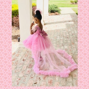 May include: A young girl wearing a pink high-low dress with a tulle train and a bow on the back. The dress has a ruffled hem and is decorated with lace.