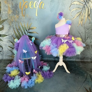 May include: A purple tulle tutu with a rainbow pom-pom trim and butterfly accents. The tutu is displayed on a white stand with a matching purple tulle veil with butterfly accents.
