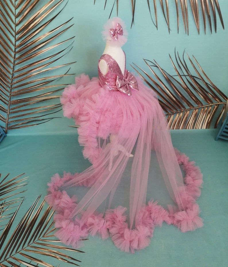 May include: A pink tulle dress with a sequined bodice and a large bow. The dress has a long train and is decorated with ruffles.