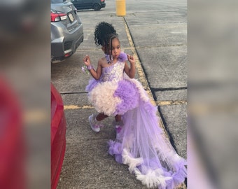 May include: A young girl wearing a lavender and white tulle dress with a high-low hemline. The dress has a beaded bodice and a ruffled skirt. The girl is wearing white trainers and has her hair in braids.
