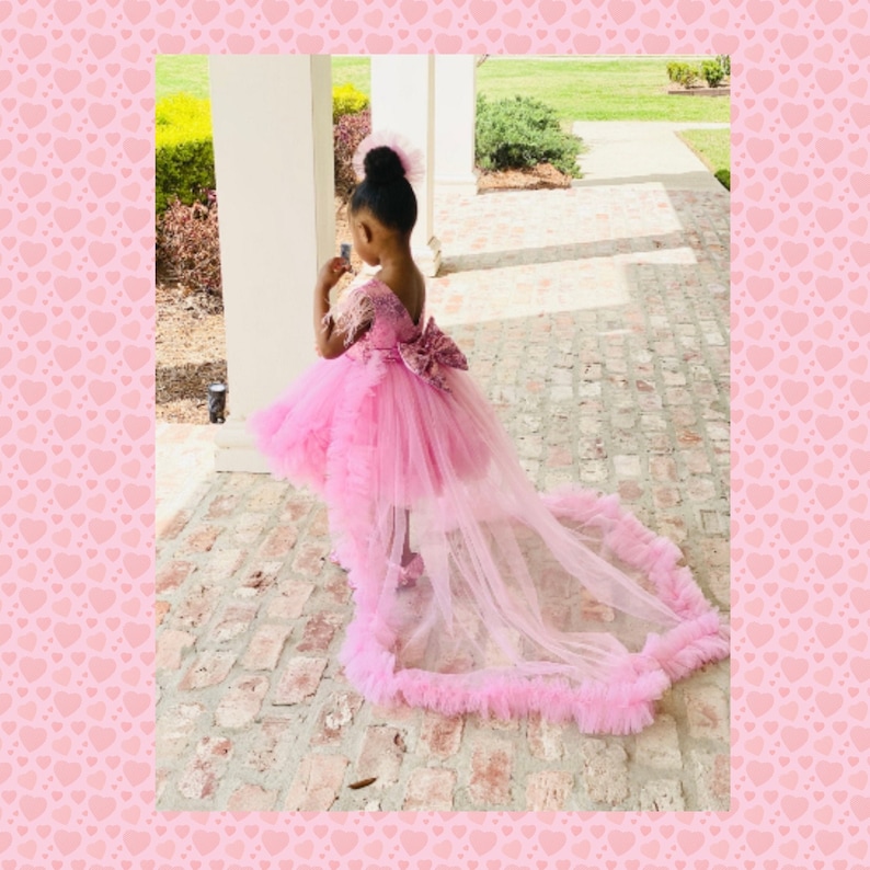 May include: A young girl wearing a pink high-low dress with a tulle train and a bow on the back. The dress has a ruffled hem and is decorated with lace.