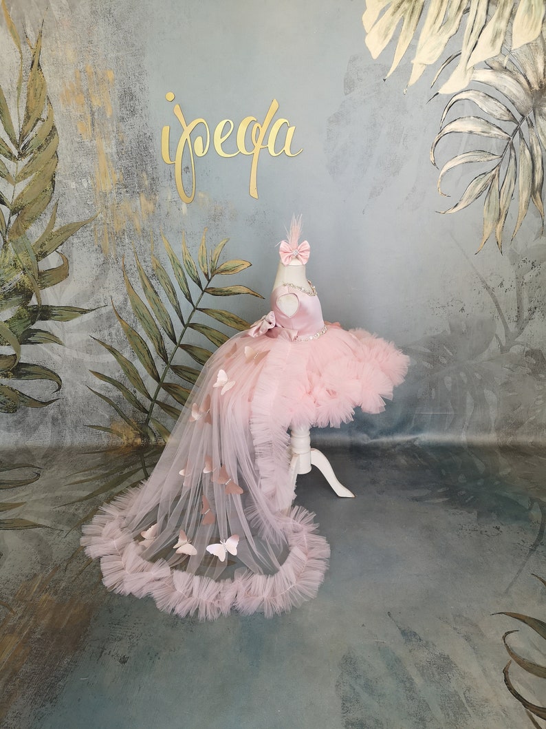 May include: A pink tulle dress with a butterfly design and a train. The dress has a sweetheart neckline and a fitted bodice. The train is adorned with butterflies and has a ruffled edge.