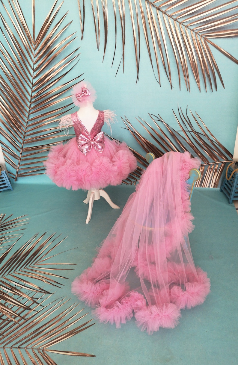 May include: A pink tulle dress with sequins and a large pink bow. The dress is on a white stand and has a matching pink tulle veil draped over a chair.