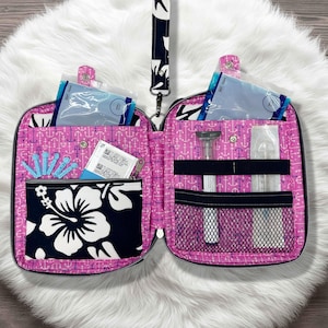 以下が含まれることがあります： 黒いジッパーが付いた、開いたピンク色の医療用オーガナイザー。ハイビスカスの花のポケットとアンカーパターンが特徴です。注射器や小さなポーチなど、医療用品が入った複数のコンパートメントがあります。白黒模様のリストストラップが付いています。