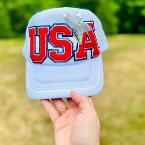May include: A light blue trucker hat with a red, white, and blue "USA" embroidered on the front. A silver sequined star is above the text.