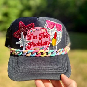 Peut inclure: Casquette de camionneur noire avec un patch rose, rouge et blanc qui dit "Ce n'est pas mon problème, c'est moi". La casquette a également une étoile argentée, une étoile rose, une boule disco et un patch de rouge à lèvres rouge.