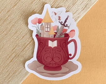 May include: A red teacup with a white saucer, filled with a miniature castle, a sword, and a book. The castle is yellow with a brown roof and a white window. The sword is silver and the book is open. There are pink flowers around the cup.