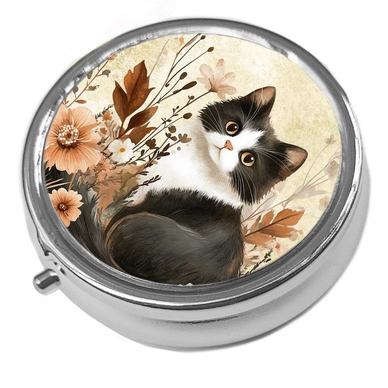 Puede incluir: Un pastillero redondo y plateado con tapa abisagrada. La tapa presenta una ilustraci&oacute;n a la acuarela de un gato blanco y negro rodeado de flores y follaje en tonos melocot&oacute;n, marr&oacute;n y crema. El gato tiene ojos amarillos.