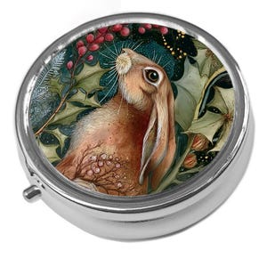 May include: A circular silver pill box featuring a detailed illustration of a brown hare amidst holly leaves and red berries. The artwork is set against a dark teal background, creating a festive and nature-inspired design.
