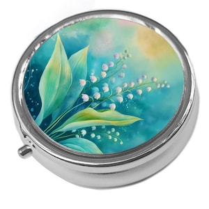 May include: A circular, silver pill box featuring a watercolour design of lily of the valley flowers and leaves. The artwork uses shades of green, blue, and yellow, creating a soft, floral aesthetic. The pill box has a small, silver clasp.