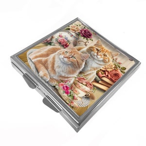 May include: A silver compact mirror featuring a colourful image of two orange tabby cats sitting on a floral patterned surface with teacups and books. The image has a vintage aesthetic.