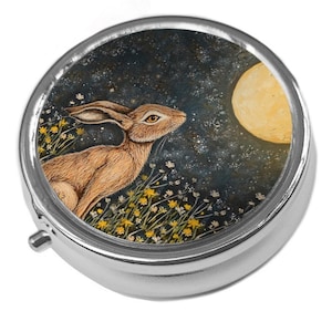 May include: A circular, silver pill box featuring a painting of a brown hare looking at a full moon against a starry night backdrop. The hare is amidst yellow and white wildflowers.