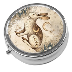 May include: A round silver pill box featuring a detailed illustration of a rabbit on the lid. The rabbit is depicted in brown and cream tones, surrounded by celestial motifs including stars, a crescent moon, and planetary phases on a beige background.