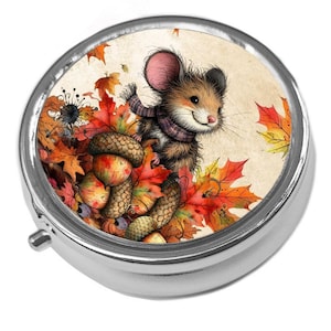 May include: A silver pill box featuring a charming illustration of a mouse in a scarf, amidst autumn leaves and acorns. The mouse is smiling, and the design has a warm feel. The pill box is round with a hinged lid, perfect for storing small items.