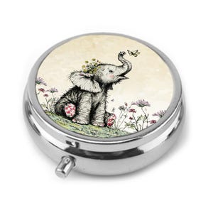 May include: A round, silver-toned metal pill box featuring a circular inset illustration of a small elephant with flowers on its head, looking up at a butterfly. The elephant is surrounded by delicate line-drawn flowers on a cream-coloured background.