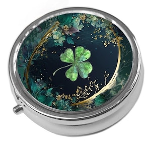 May include: A silver pill box featuring a four-leaf clover design. The clover is green, set against a dark blue backdrop with green and gold floral details. The pill box has a hinged lid and a small opening tab.