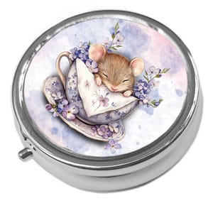 May include: A circular, silver pill box with a charming design. The lid displays a watercolour illustration of a sleeping mouse in a teacup surrounded by purple flowers. The teacup and saucer have a floral pattern.