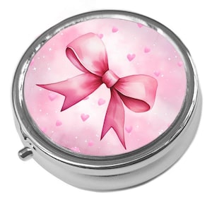 May include: A silver pill box featuring a pink bow design. The bow is a watercolour illustration with a gradient of pink hues. The background is a soft pink with small pink hearts and white specks.