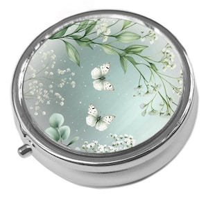 May include: A silver pill box with a floral and butterfly design. The lid features a light blue background with white butterflies, green leaves, and delicate white flowers. The pill box is round and has a small, silver clasp.