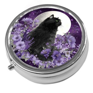 May include: A round silver pill box featuring a printed image of a fluffy black cat with green eyes sitting among purple flowers against a backdrop of a large white full moon and a dark purple starry night sky. The metal case has a hinged lid.