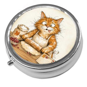 May include: A round silver metal pill box featuring a colourful illustration of a fluffy orange cat wearing an apron, using a wooden rolling pin to flatten dough on a wooden surface, with a small bag of flour, red berries, and a recipe card nearby.