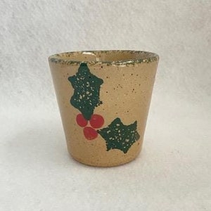 May include: A small, beige ceramic cup with a festive holly design. The cup features green holly leaves and red berries, perfect for holiday decor. The cup is approximately 3 inches tall.