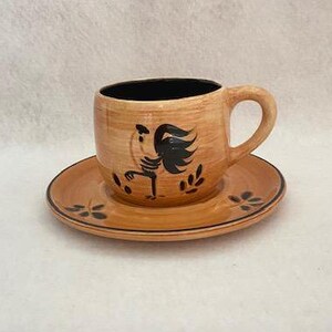 May include: A brown ceramic coffee cup with a black rooster design and a matching saucer. The cup has a black rim and a black handle.