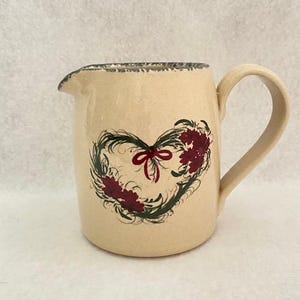 May include: Cream-colored ceramic pitcher with a heart-shaped floral design. The heart is composed of green leaves, burgundy flowers, and a red bow. The rim has a dark gray, textured edge, and the handle is curved.