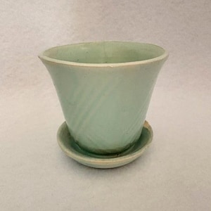May include: A pale mint green ceramic plant pot with a flared rim and a matching circular saucer. The exterior of the pot features a subtle diagonal ribbed texture, and the base shows a natural, unglazed clay edge where it meets the saucer.
