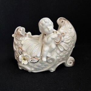May include: A white ceramic planter featuring a cherub playing a harp. The planter has a shell-like design with gold accents on the edges, flowers, and leaves. The cherub is seated inside the planter.