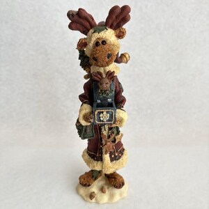 May include: A decorative figurine of a moose wearing a burgundy coat with cream-colored trim and a small moose in a carrier. The moose has brown antlers and is standing on a snow-like base. A festive holiday decoration.