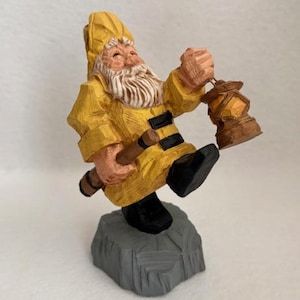 May include: A carved wooden figurine of a smiling gnome wearing a yellow coat and hat, black boots, and holding a lantern. The gnome is perched on a gray rock base, with a wooden staff in his other hand.