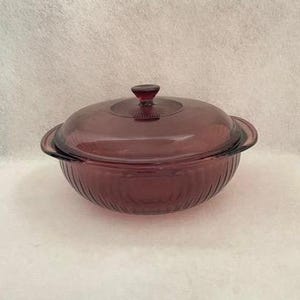 May include: A round, deep purple glass casserole dish with a matching lid. The bowl and lid feature a vertical ribbed texture along the sides. The lid includes a small, circular knob handle at the center and two integrated side handles.