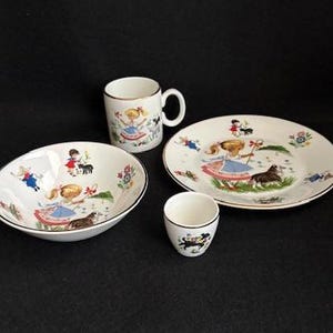 May include: A white ceramic children's dish set with a whimsical illustration of a girl, a dog, and other figures. The set includes a bowl, a plate, a mug, and a small cup. The edges are trimmed in black.