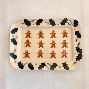 May include: Rectangular ceramic serving tray with a cream-colored background. The tray features a border of dark green holly leaves and red berries. The center has a pattern of twelve gingerbread men.