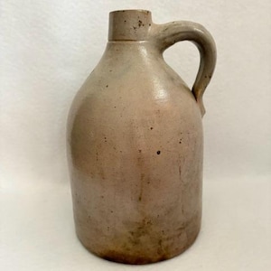 May include: A tall, cylindrical stoneware jug with a light beige, speckled glaze. The vessel features a narrow neck, a thick, curved handle attached to the side, and a rounded body with subtle variations in color and texture across the surface.