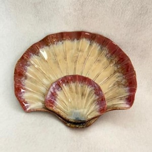 May include: A ceramic seashell-shaped dish with a fan-like design. The dish features a cream-colored interior with a reddish-brown rim and accents. The smaller shell is attached to the larger one, creating a layered effect. The dish is likely used for decorative or serving purposes.