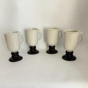 May include: A set of four ceramic mugs, each featuring a cream-colored cylindrical body with a handle and a dark brown, rounded pedestal base. The mugs are arranged in a row, showcasing a two-tone design with a smooth, glossy finish throughout.