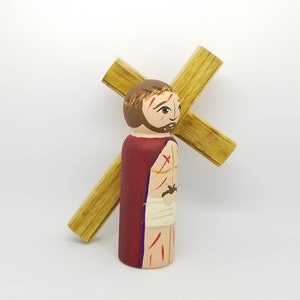 May include: A wooden figurine of Jesus carrying a wooden cross. The figurine is painted with realistic details, including a red robe, a crown of thorns, and wounds on his body. The cross is painted brown and has a simple design.