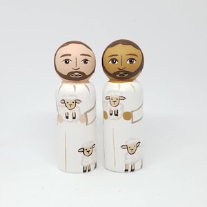 May include: Two wooden peg dolls, each with a painted face and beard. They wear white robes and hold a sheep. Additional sheep are painted on the robes. The dolls have different skin tones.
